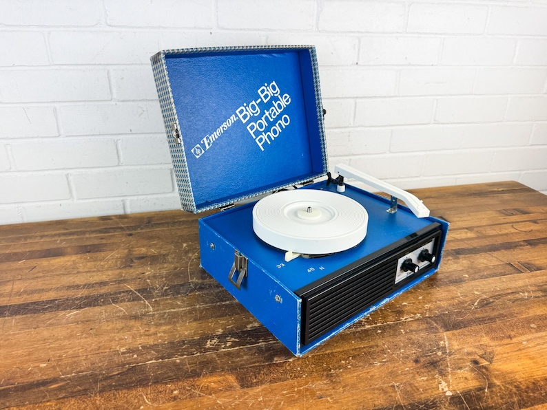 May include: A vintage Emerson Big-Big Portable Phono record player. The blue vinyl-covered case is open, revealing a white turntable and arm. The front has black speaker vents and control knobs. The case is on a wooden surface.