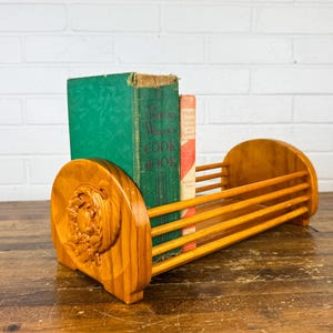 May include: A wooden book holder with a carved design on the side, holding two vintage cookbooks. The holder is made of light-colored wood and has a slatted design. The books are green and red.
