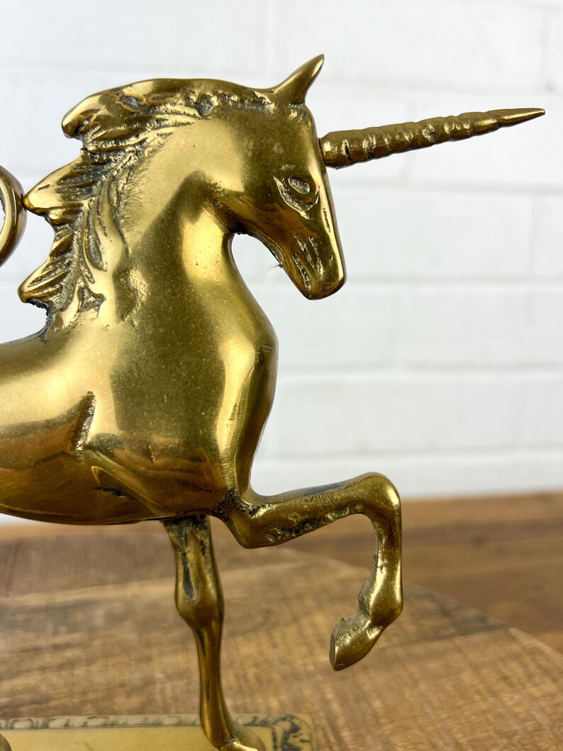 Puede incluir: Una figurita de unicornio de lat&oacute;n dorado con una melena y un cuerno detallados. El unicornio est&aacute; de pie sobre una base con un dise&ntilde;o decorativo.