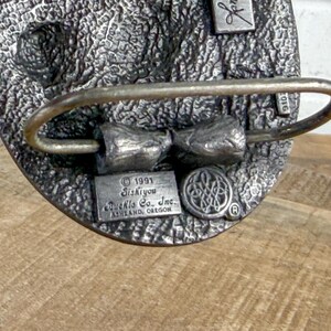 May include: A silver-toned metal belt buckle with a textured surface. The buckle features a bow-tie design and the text "1991 Siskiyou Buckle Co. Inc. Ashland, Oregon". A small emblem and the number "2016" are also visible.