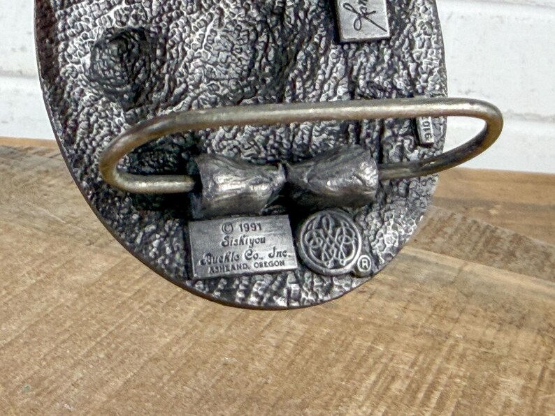 May include: A silver-toned metal belt buckle with a textured surface. The buckle features a bow-tie design and the text "1991 Siskiyou Buckle Co. Inc. Ashland, Oregon". A small emblem and the number "2016" are also visible.