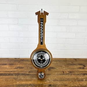 May include: A wall-mounted weather station featuring a wooden frame with a vertical thermometer, a large central barometer with a black dial, and a smaller hygrometer at the bottom. The thermometer displays both Celsius and Fahrenheit scales.
