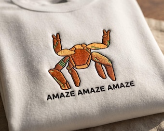 May include: A cream-colored crewneck sweatshirt featuring an embroidered graphic of a brown crab with green accents, making a peace sign with its top claws. Below the crab, the text AMAZE AMAZE AMAZE is embroidered in black thread.