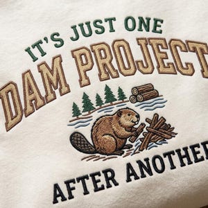 Puede incluir: Una sudadera de color crema con un diseño bordado de un castor marrón construyendo una presa con troncos. El texto dice IT'S JUST ONE DAM PROJECT AFTER ANOTHER en letras de bloque verde oscuro y marrón, junto a pinos verdes.