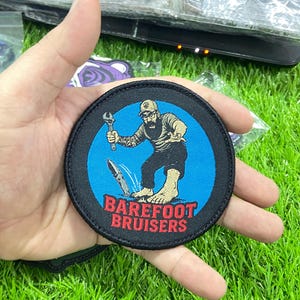 Könnte beinhalten: Ein kreisförmiger bestickter Aufnäher mit schwarzem Rand und leuchtend blauem Zentrum. Das Motiv zeigt eine bärtige Figur mit Kappe, die einen Schraubenschlüssel hält und barfuß neben einem Messer steht. Der Text BAREFOOT BRUISERS ist rot.