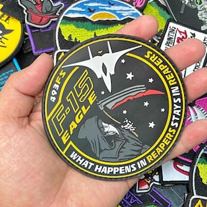 May include: A circular black patch featuring a white F-15 Eagle jet silhouette, a grim reaper figure holding a scythe, and white stars. The text reads 493FS F-15 EAGLE and WHAT HAPPENS IN REAPERS STAYS IN REAPERS in yellow and white lettering.