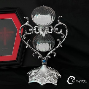 May include: A silver-toned hourglass featuring a clear glass bulb filled with white sand. The frame has an ornate, curved design with a heart-shaped center piece and a wide, textured base adorned with floral patterns and a small blue circular accent.