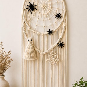 May include: A macrame wall hanging featuring a circular frame with a woven web pattern, adorned with four small black spiders, a crocheted ghost figure with black eyes, and a small crocheted spiderweb motif, all finished with long hanging fringe.