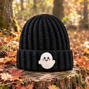 May include: A black ribbed knit beanie featuring a small, white crocheted ghost appliqué on the front cuff. The ghost has two black bead eyes, a small black stitched mouth, and two tiny pink circular cheeks, creating a simple, friendly expression.