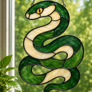 Könnte beinhalten: Ein Buntglas-Suncatcher in Form einer gewundenen Schlange mit dunkelgrünen und cremefarbenen, strukturierten Glasscheiben. Die Schlange hat ein markantes gelbes Auge mit schwarzer Pupille, eingefasst in dicke schwarze Metallstege, an einer Kette.