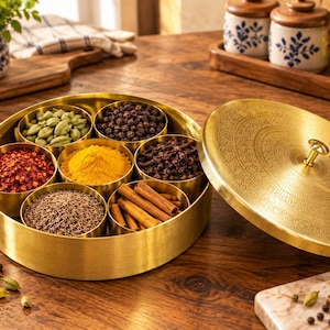 Könnte beinhalten: Eine runde Gewürzdose aus Messing mit sieben herausnehmbaren Inneneinsätzen, gefüllt mit verschiedenen Gewürzen wie Kardamom, schwarzem Pfeffer, Chiliflocken, Kurkuma, Nelken, Kreuzkümmel und Zimtstangen, inklusive eines passenden gravierten Deckels.