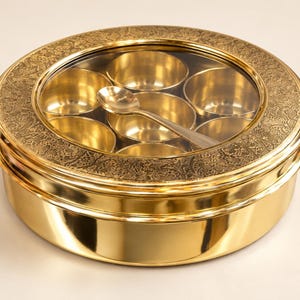 Op de afbeelding: Een ronde kruidendoos van messing met een gepolijste afwerking en een helder glazen deksel. Het deksel heeft een ingewikkelde gegraveerde bloemenrand. Binnenin staan zeven kleine messing kommetjes in een cirkel, met een messing lepel.