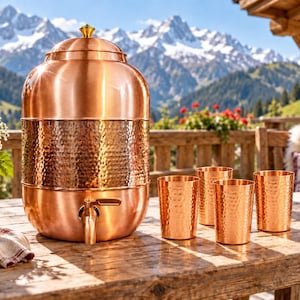 Könnte beinhalten: Ein großer Wasserspender aus poliertem Kupfer mit einem gehämmerten Metallband in der Mitte und einem passenden Zapfhahn, begleitet von vier passenden gehämmerten Kupferbechern, alles auf einer rustikalen Holzoberfläche im Freien.