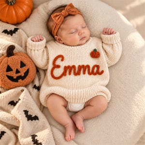 Può includere: Un maglione in maglia a maniche lunghe color crema con il nome Emma ricamato in filo arancione bruciato sul petto, impreziosito da una piccola applicazione a forma di zucca, abbinato a una fascia con fiocco coordinata per neonati.