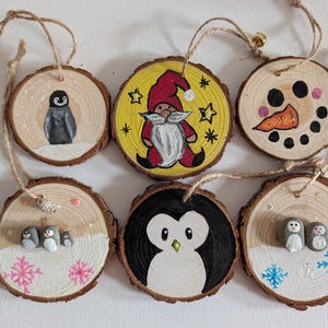 May include: Six round wooden slice ornaments with bark edges, each featuring hand-painted winter designs including a penguin, a gnome on a yellow background, a snowman face, and small pebble-based penguin figures with snowflakes.