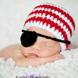 May include: A sleeping baby wearing a red and white striped crocheted hat with a black eye patch. The baby is lying on a white fluffy blanket.