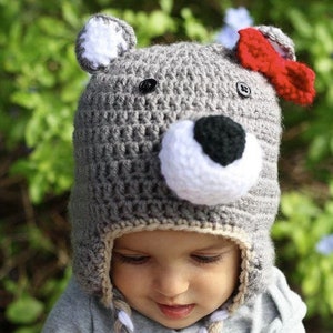 Puede incluir: Un gorro de oso de punto gris con detalles blancos y negros y un lazo rojo. El gorro tiene un cordón trenzado largo con hilo blanco, gris y beige.