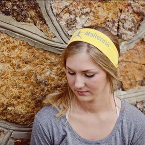May include: A young woman with blonde hair is wearing a yellow headband with the name "Makenna" written on it. She is wearing a gray shirt and is standing in front of a stone wall.