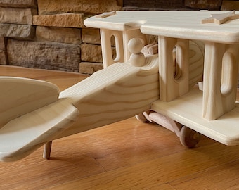 May include: A wooden toy airplane with a natural finish. The biplane features a pilot figure, two wings, a tail, and wheels. The toy is made of light-colored wood and is designed for children.