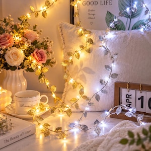 May include: A string of decorative fairy lights featuring metallic gold and silver leaf-shaped accents. The lights emit a warm yellow and cool white glow, intertwined with a white pillow, a ceramic cup, and a floral arrangement in a white vase.