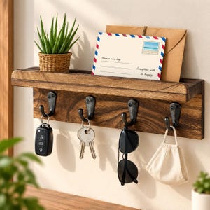 May include: A dark wood wall-mounted organizer shelf featuring four black metal hooks. The shelf holds a small green plant in a woven pot and a postcard with the text "Have a nice trip" and "Everytime with to happy." Items hanging include keys, sunglasses, and a mask.