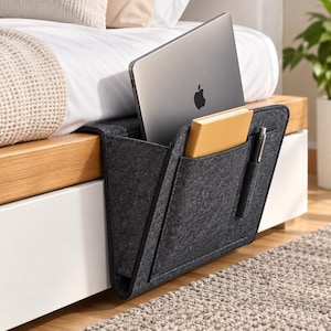 May include: A dark gray felt bedside storage caddy featuring a large main compartment holding a silver laptop, a smaller front pocket containing a tan notebook, and a narrow side slot holding a silver pen, designed to hang over a bed frame edge.