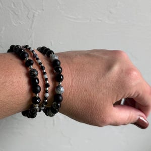 May include: A set of three beaded bracelets featuring a combination of matte black, glossy black, and grey speckled beads. The design incorporates small silver-toned metallic spacers and clear crystal accents, creating a layered look with varying bead sizes and textures throughout the three strands.
