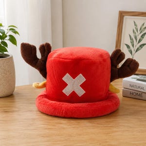 May include: A plush red bucket hat featuring two large brown antlers extending from the sides and a white cross emblem stitched onto the front. The hat is made of a soft, fuzzy material and has a wide, rounded brim, mimicking a character design.