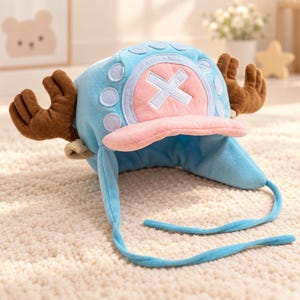 May include: A light blue plush hat featuring a pink circular patch with a white X symbol on the front. The hat includes two brown plush antlers extending from the sides, a pink brim, and two long blue fabric ties hanging from the ear flaps.