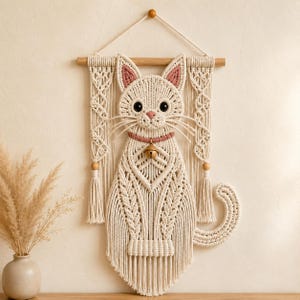 May include: A macrame wall hanging featuring a cream-colored cat design with pink inner ears and a pink collar with a small gold bell. The cat is suspended from a wooden dowel with decorative side panels, fringe, and a curved tail detail.