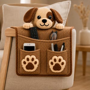 May include: A brown knitted armchair organizer featuring a plush dog head at the top and two front pockets decorated with cream and brown paw prints. The organizer holds a remote control, eyeglasses, a smartphone, and three pens.