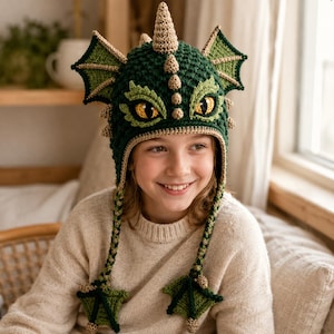 May include: A dark green crocheted hat designed to resemble a dragon, featuring large pointed ears, a central row of small beige spikes, and two yellow eyes with black pupils. The hat includes long braided earflaps ending in small green dragon feet.