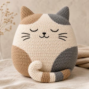 May include: A round, stuffed crochet cat toy featuring a calico pattern with patches of cream, tan, and dark grey yarn. The cat has embroidered closed eyes, a small nose, and whiskers, with a curled tail resting at the front of its body.