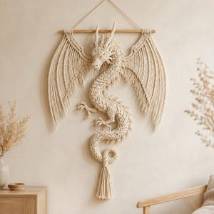Puede incluir: Un gran tapiz de macramé en color blanco roto con forma de dragón, que presenta intrincados patrones de nudos, alas extendidas y una larga cola curva que termina en una borla. La pieza cuelga de un soporte de madera horizontal.