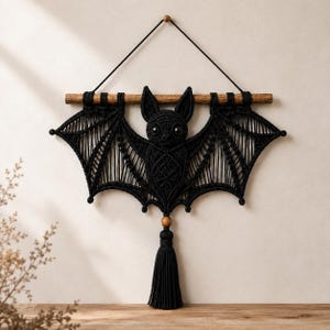 May include: A black macrame wall hanging shaped like a bat, suspended from a horizontal wooden branch by a black cord. The bat features intricate knotted patterns, two pointed ears, and two round black eyes, with a wooden bead and tassel below.