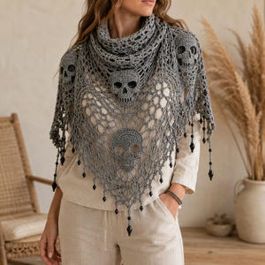 Peut inclure: Un châle en crochet gris présentant un motif ajouré complexe avec trois motifs de crânes intégrés au design. Le vêtement triangulaire est agrémenté de perles noires pendantes le long du bord inférieur et sur les côtés.
