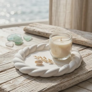 May include: A round white tray with a braided rope-style rim. A clear glass candle holder containing a cream-colored wax candle sits on the tray, accompanied by a small beige seashell and a single dried stalk of wheat resting on the flat surface.