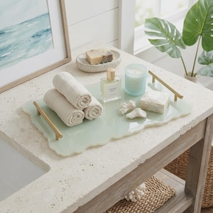 May include: A rectangular light green resin tray with wavy edges and two gold-toned metal handles. The tray holds three rolled beige cotton towels, a glass perfume bottle labeled Oceanic Drift, a white candle in a glass, and two white coral pieces.