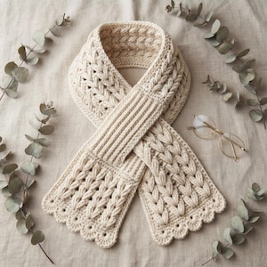 May include: A cream-colored crocheted scarf featuring a textured cable knit pattern, a ribbed center section, and scalloped edges. The scarf is displayed alongside a pair of thin-rimmed glasses with clear lenses, arranged on a neutral fabric surface.