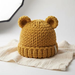 May include: A mustard yellow beanie crafted from thick, plush yarn featuring two rounded bear-like ears on top. The hat has a ribbed cuff at the base and a small white fabric tag attached to the side, showcasing a textured, chunky knit construction.