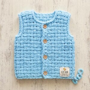 May include: A light blue sleeveless vest made of thick, plush yarn with a woven basket-weave texture. The front features four round wooden buttons and a white fabric label at the bottom right corner with the text ELIF HOBI and a bird icon.