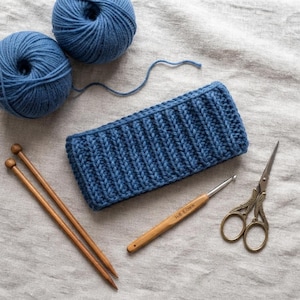 Puede incluir: Una diadema tejida azul rectangular con textura acanalada, dos ovillos de lana azul, dos agujas de tejer de madera, un ganchillo de madera marcado H/8 5.0mm y unas tijeras de metal ornamentadas con detalles intrincados en el mango.