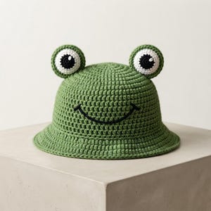 Op de afbeelding: Een groene gehaakte bucket hat met twee uitstekende kikkerogen met witte en zwarte details. De hoed heeft een gestikte zwarte gebogen lijn die een eenvoudige glimlach op de voorkant vormt. De textuur is gemaakt van dichte lussen van garen.