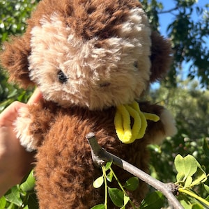 May include: A plush monkey toy with brown and cream-colored fur. The stuffed animal features a round head, small ears, and a bright yellow fuzzy ribbon tied around its neck. The toy has dark, round eyes and a soft, textured appearance throughout.