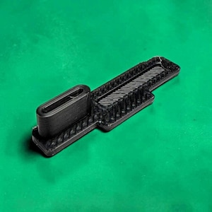 May include: A black 3D-printed plastic component featuring a rectangular base with a textured, grid-like surface pattern. Attached to one end is a raised, hollow, oval-shaped vertical loop with visible horizontal layer lines from the printing process.