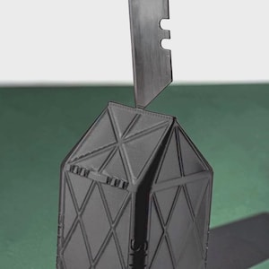 May include: A black, three-dimensional, geometric house-shaped object with a diamond-patterned exterior. A single, thin, metallic razor blade is inserted vertically into the top peak of the structure, extending upwards from the roof.