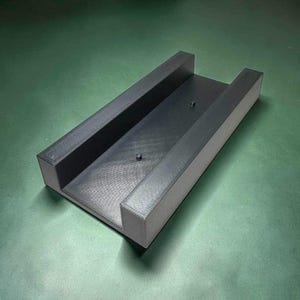 May include: A rectangular black tray with raised side walls and two small, centred circular pegs protruding from the flat base. The object features a textured, matte finish and an open-ended design on both ends of the structure.