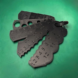 以下が含まれることがあります： 黒いリングでまとめられた、3Dプリント製の黒いプラスチック製編み針ゲージと目数カウンターのセット。各ツールには浮き彫りの数字と、針の直径を測定したり目を数えたりするためのさまざまなサイズの穴や切り込みが施されています。