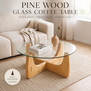 May include: A round coffee table with a clear glass top and a light-coloured solid pine wood base featuring a cross-shaped design. The table holds two stacked books and a small beige ceramic vase. Text on the image reads PINE WOOD GLASS COFFEE TABLE.