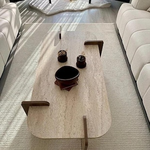 May include: An oval coffee table with a beige travertine stone top and dark brown wooden legs that wrap around the edges. The table surface holds a dark brown bowl, two small dark brown pots with dried plants, and a set of square coasters.